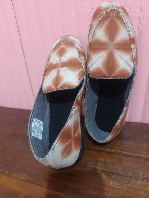 Sepasang sepatu slip on bermotif shibori cokelat putih di atas meja kayu berlatar dinding merah muda