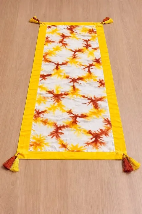 Taplak meja panjang bermotif shibori jingga di atas kain putih, bertepi kuning dengan rumbai di sudutnya