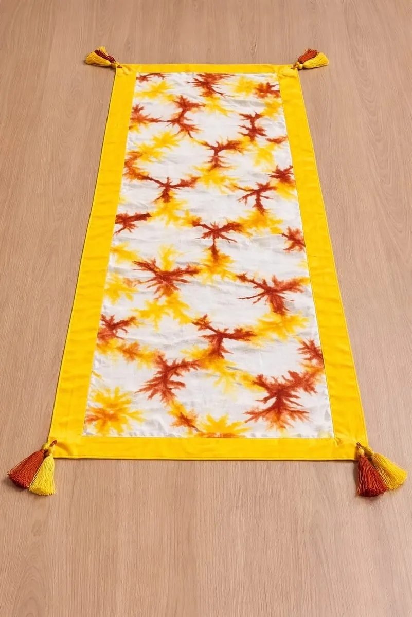 Taplak meja panjang bermotif shibori jingga di atas kain putih, bertepi kuning dengan rumbai di sudutnya