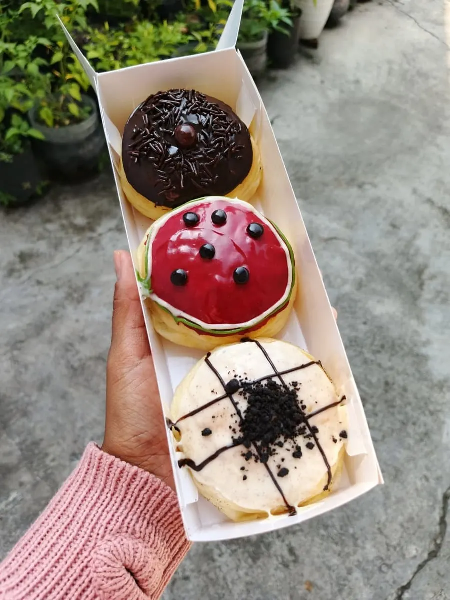 Tangan memegang kotak putih berisi tiga donat bertopping cokelat, glasir merah dan krim putih dengan remahan biskuit