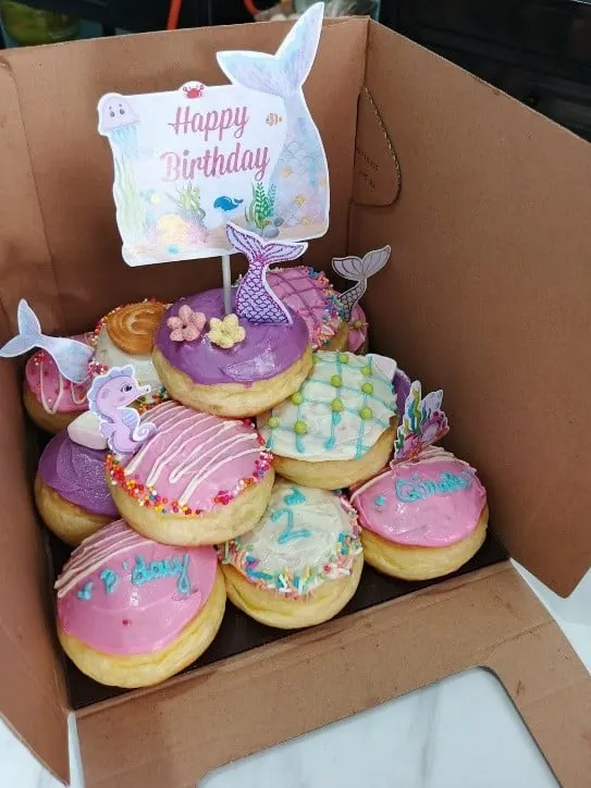 Kotak berisi tumpukan donat glasir ungu, merah muda dan putih berhias ekor duyung dan tulisan Happy Birthday