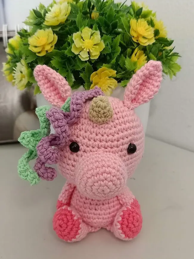 Boneka rajut unicorn merah muda dengan surai hijau dan ungu di depan pot bunga kuning