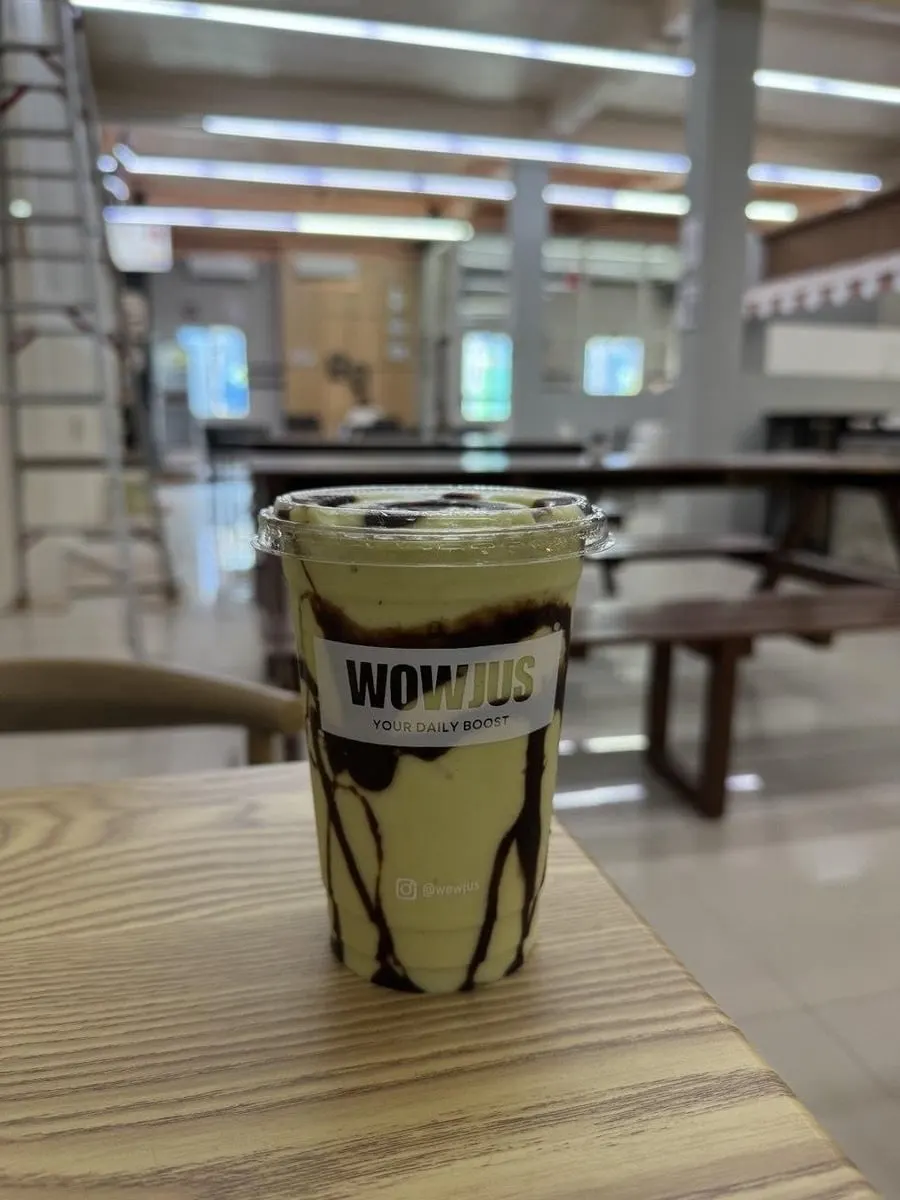Gelas WOWJUS berisi smoothie hijau muda dengan lelehan cokelat di dinding gelas, di atas meja kayu