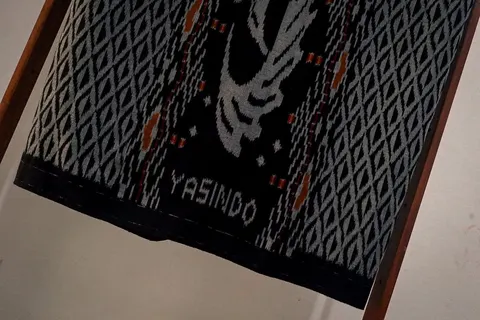 Sarung tenun ikat hitam bermotif belah ketupat abu-abu dengan tulisan Yasindo, tergantung di rangka kayu