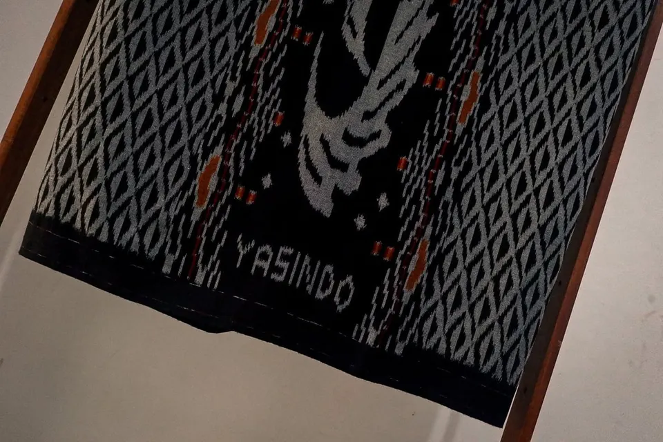 Sarung tenun ikat hitam bermotif belah ketupat abu-abu dengan tulisan Yasindo, tergantung di rangka kayu