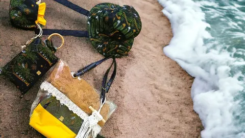 Tas kain motif hitam hijau, topi bucket dan tas transparan berisi serat cokelat dengan renda di atas pasir pantai