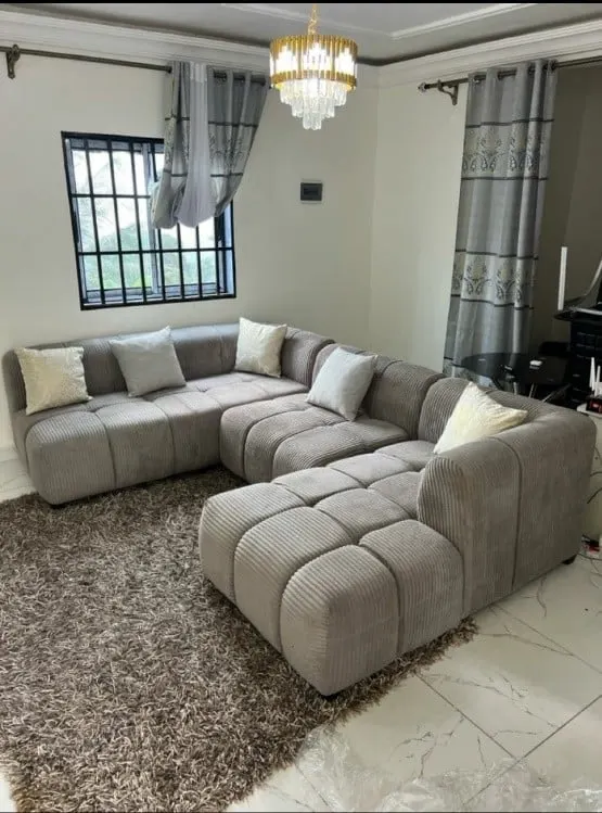 Sofa sudut modular abu-abu dengan bantal krem di ruang tamu berlantai marmer dan karpet bulu