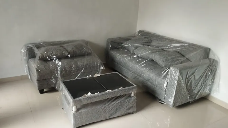 Satu set sofa abu-abu dan meja kaca hitam yang masih terbungkus plastik di ruangan putih