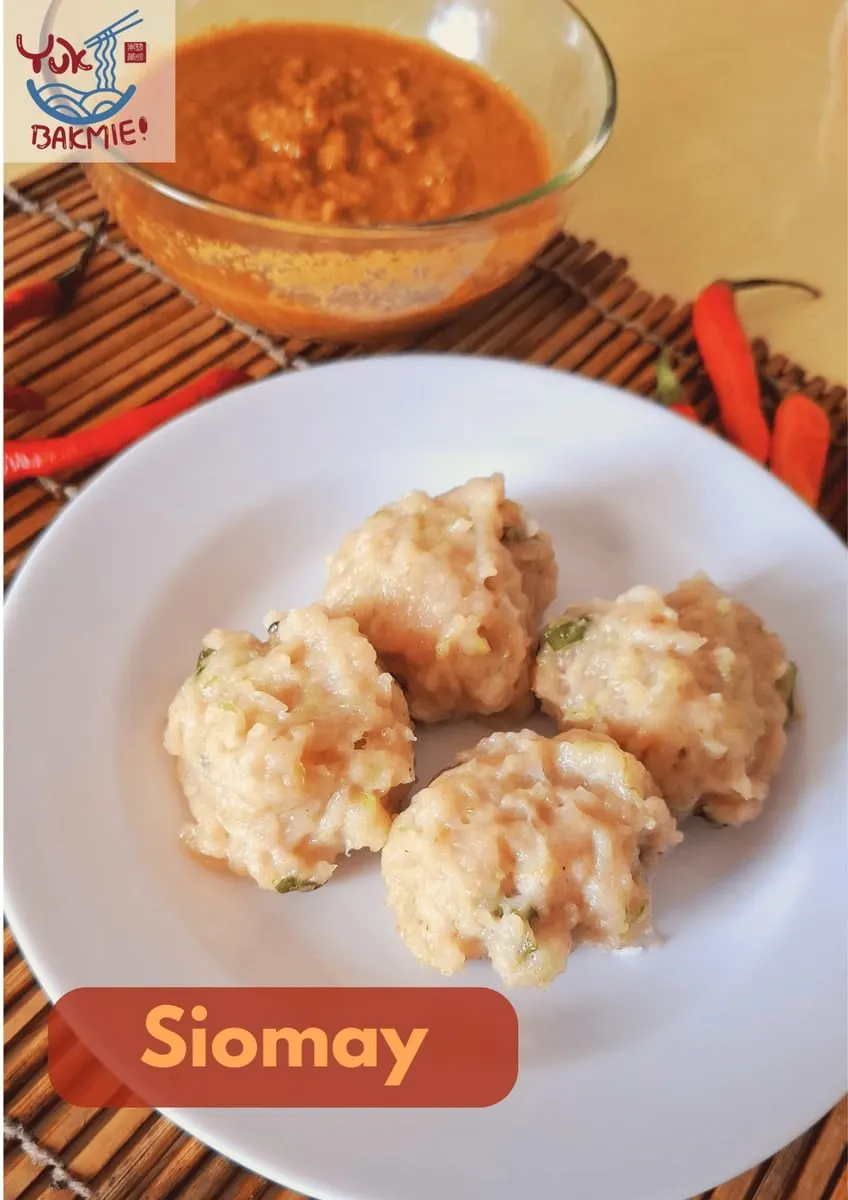 Empat siomay di piring putih dengan semangkuk saus kacang dan cabai merah di atas tikar bambu