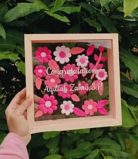Bingkai kayu merah muda berisi bunga hias merah muda dan putih dengan tulisan Congratulations dan nama penerima