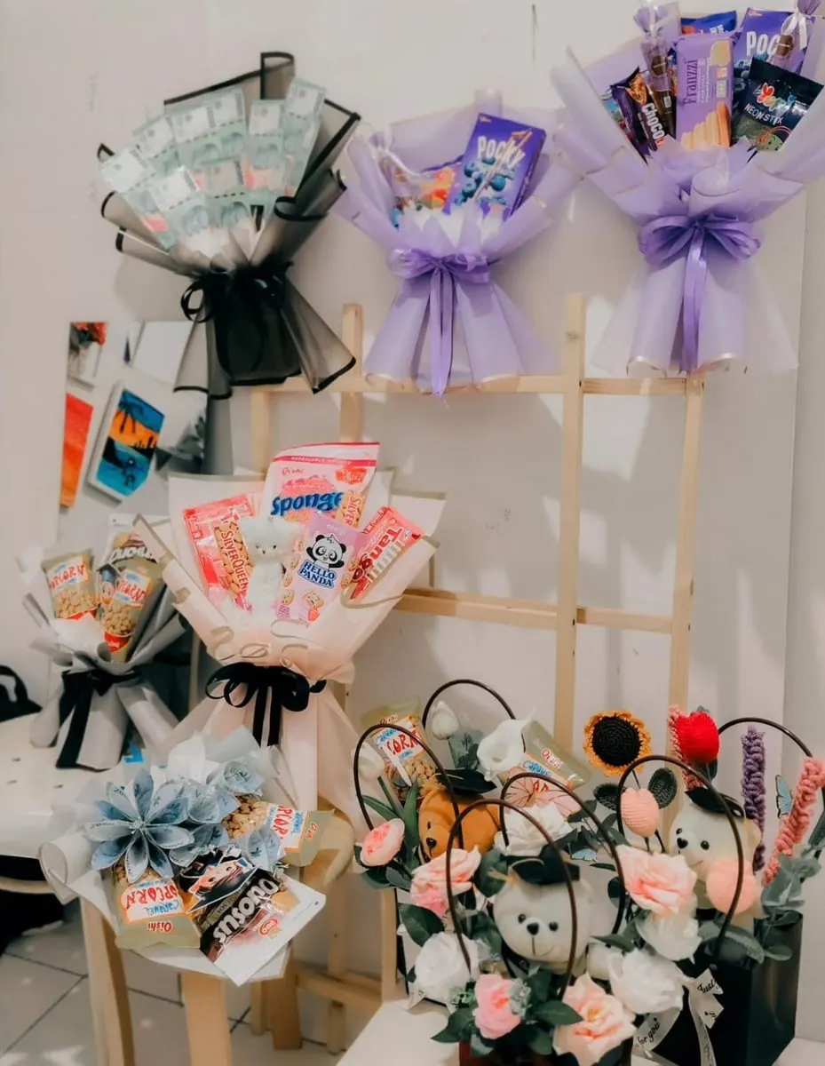 Rak kayu dengan buket uang, buket snack berbungkus ungu dan putih, serta buket boneka dan bunga rajut