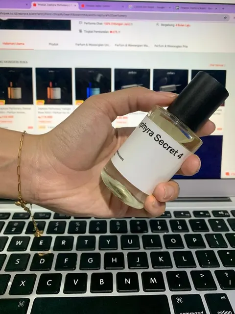 Tangan memegang botol parfum Zephyra Secret 4 bertutup hitam di depan laptop yang membuka toko daring